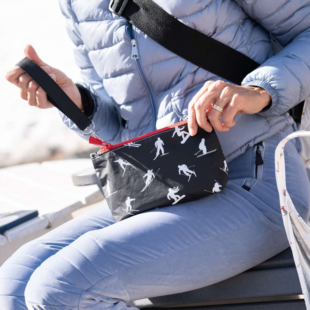 A person in a light blue puffer jacket and matching pants holds a black wristlet bag with a red zipper and a white ski silhouette pattern. The person is seated outdoors.