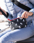 A person in a light blue puffer jacket and matching pants holds a black wristlet bag with a red zipper and a white ski silhouette pattern. The person is seated outdoors.