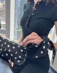 A person wearing a black short-sleeve top holds a black wristlet clutch bag featuring a white star and moon pattern with silk scarfette used as wristlet strap. The person is seated indoors near a window with a city view.