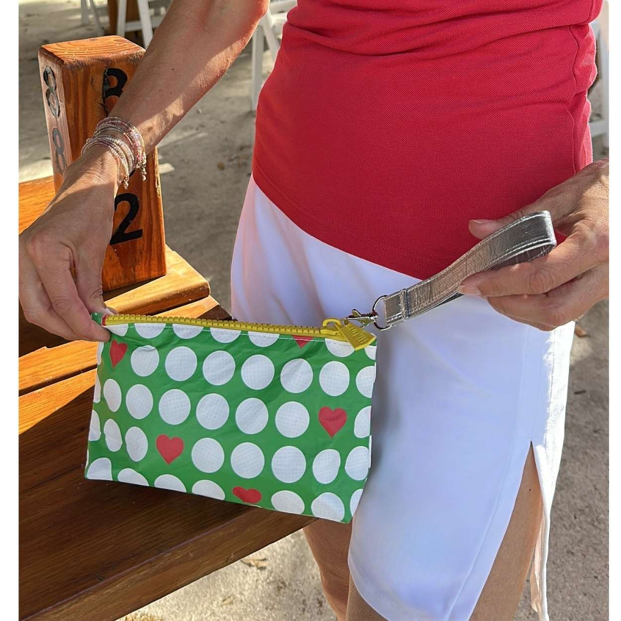 Person holding a green wristlet pouch with white golf balls and red hearts, standing outdoors.