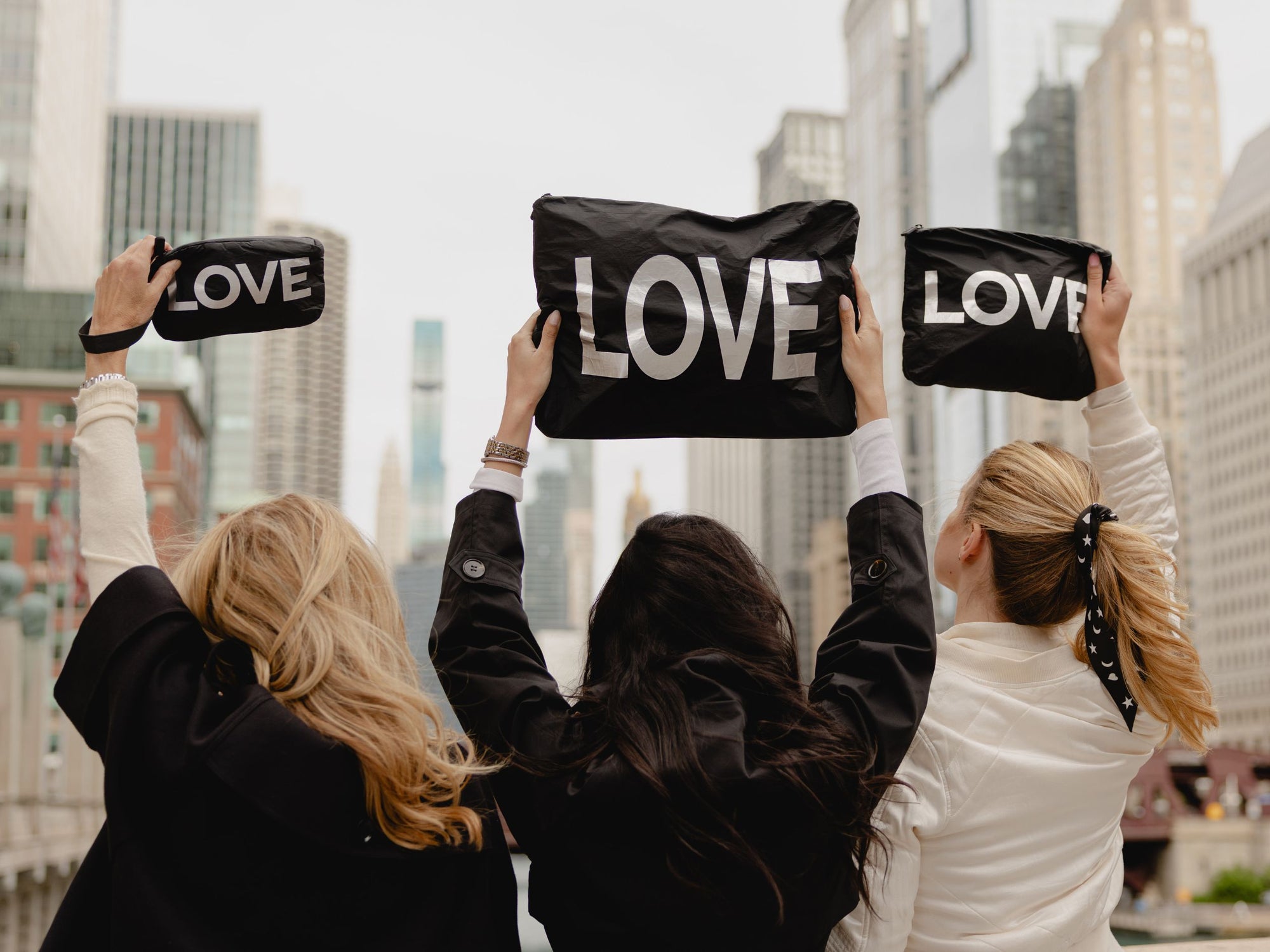 Three women seen from behind holding up black zipper pouch bags printed with the word 'LOVE' in white letters, set against a blurred Chicago city skyline background.