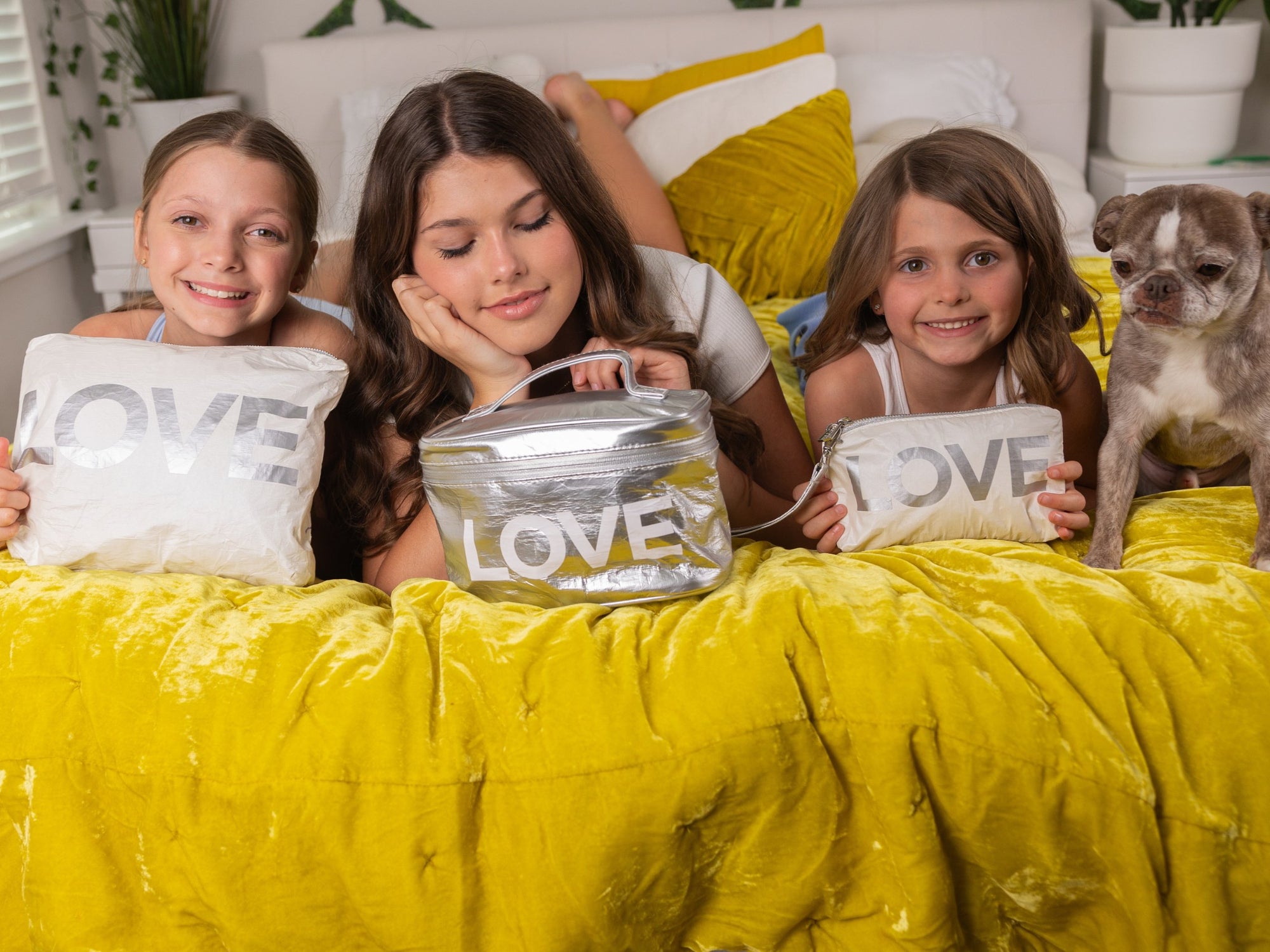 Three young girls and a small dog are lying on a bed covered with a bright yellow blanket, each holding a white pillow or bag printed with the word LOVE in large letters. The scene is indoors and styled as a cozy bedroom setting.