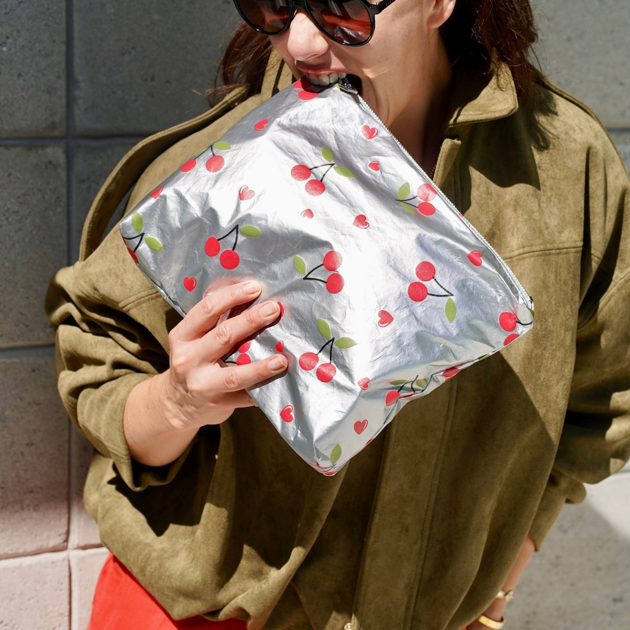 Person holding a silver pouch with red cherry pattern against a gray wall.