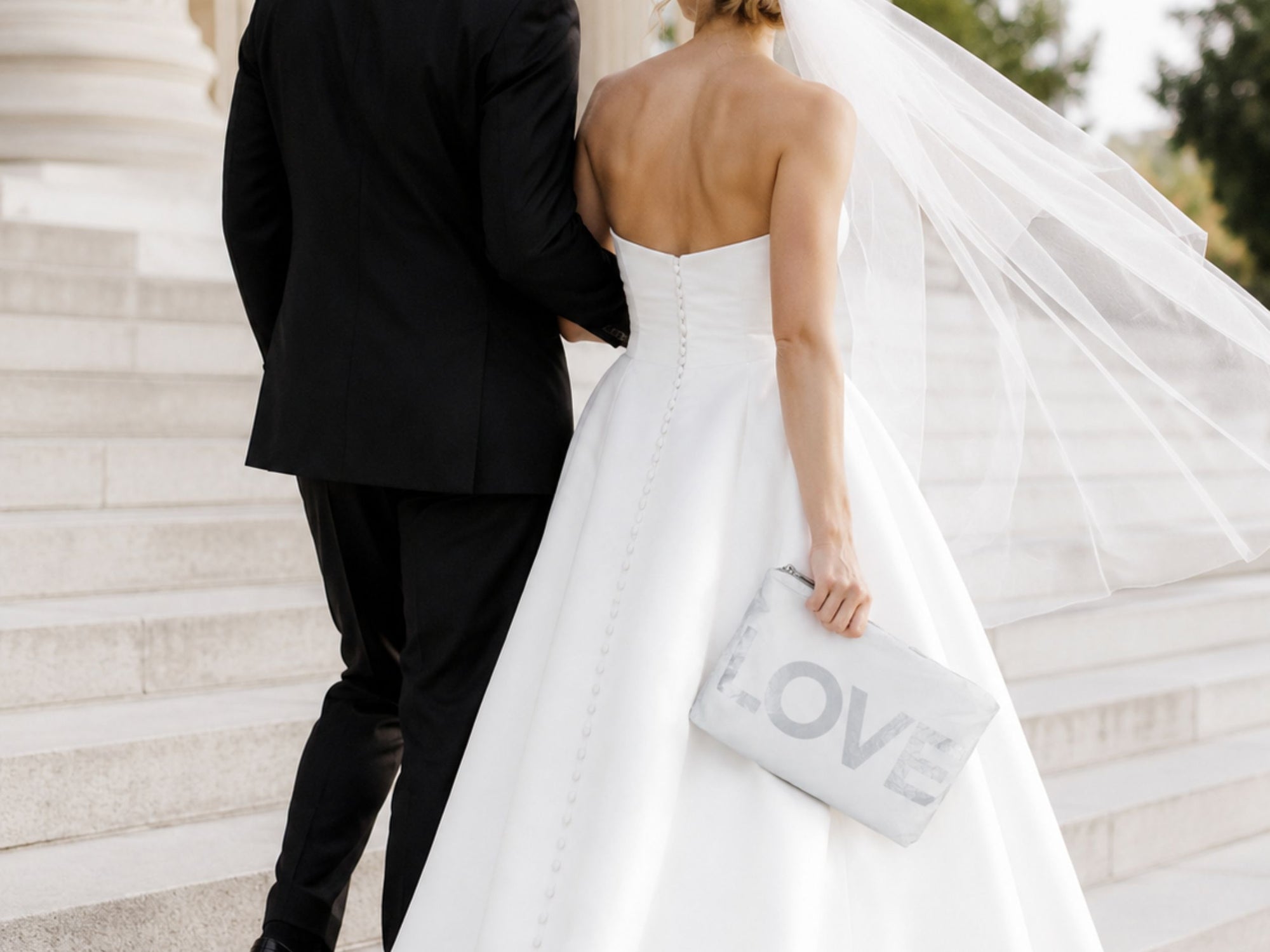 Couple in wedding attire with a 'LOVE' pouch, standing on steps.