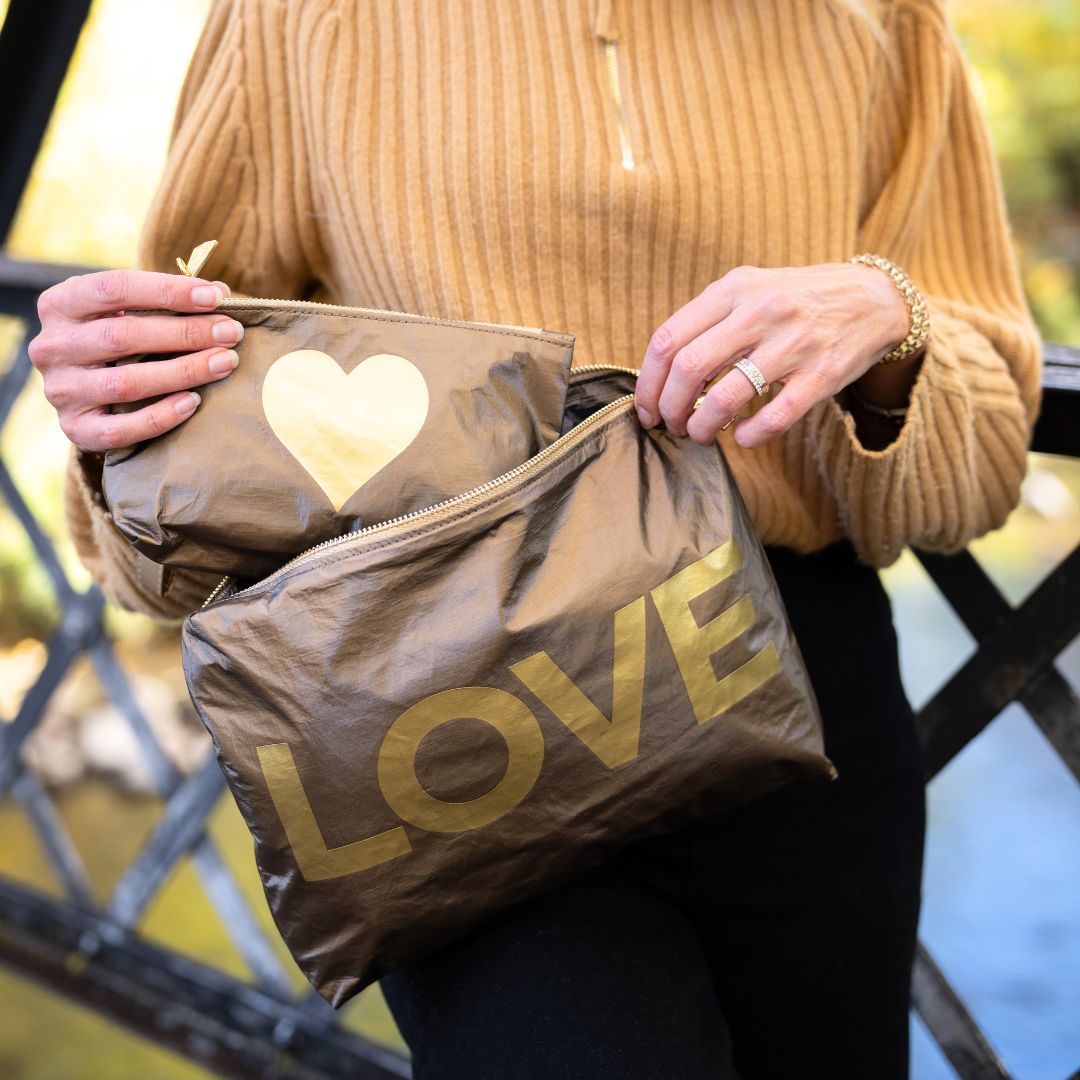 A person wearing a tan ribbed sweater holds two metallic bronze zip pouches outdoors. One pouch has a gold heart, and the larger front pouch has the word LOVE in large gold letters.