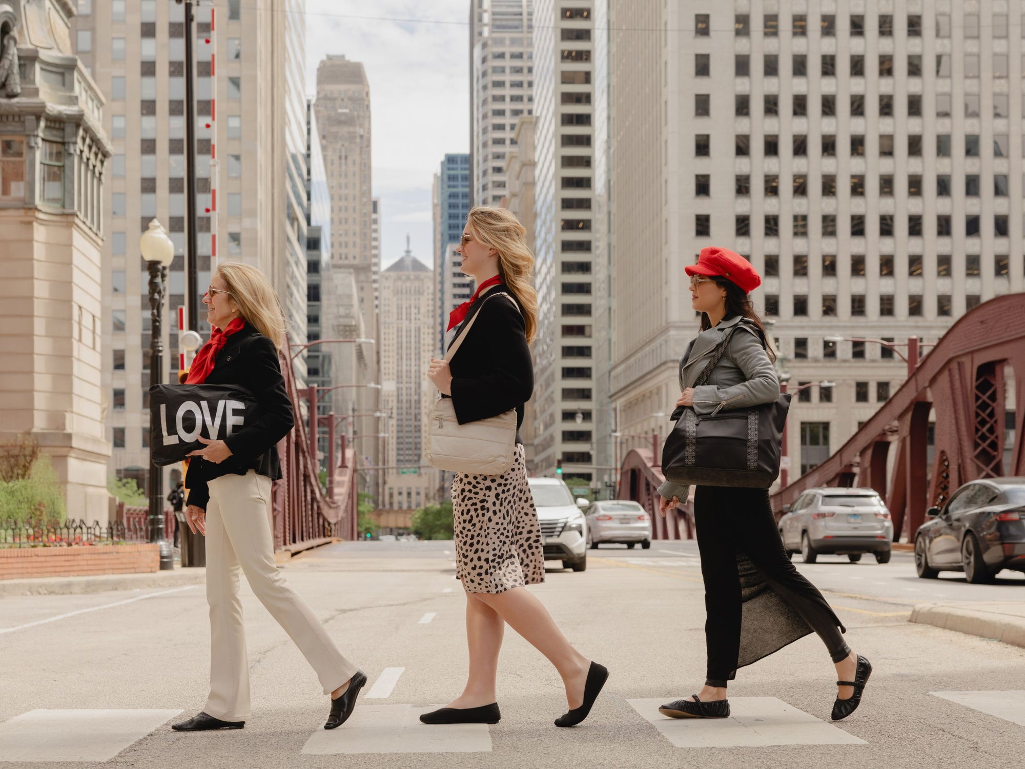 Three women crossing the street in river north Chicago. One holding a black silver LOVE zipper pouch. Another woman has a shimmer beige puffer purse on her shoulder and the 3rd girl has a black tote on her shoulder.
