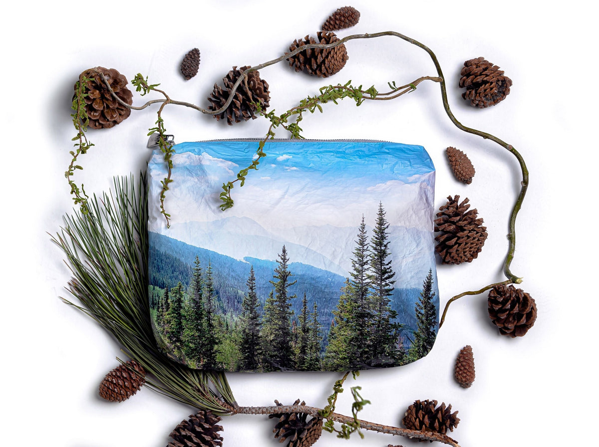 Medium zipper pouch depicting the top of a mountain with evergreen trees on white background with pinecones 