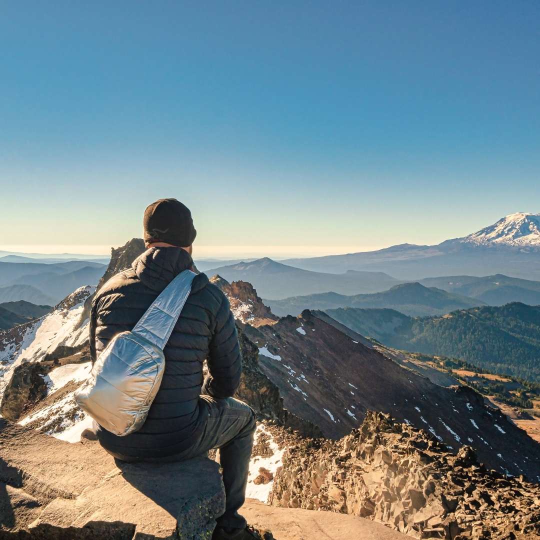 Person sitting on a mountain peak with a silver backpack, looking at a scenic view of mountains.