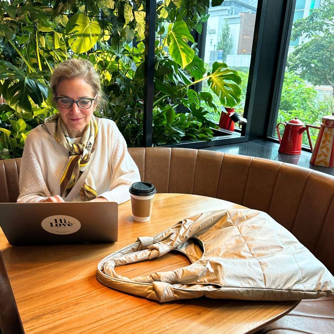 Woman working on a laptop in a cafe with plants and a beige tote bag