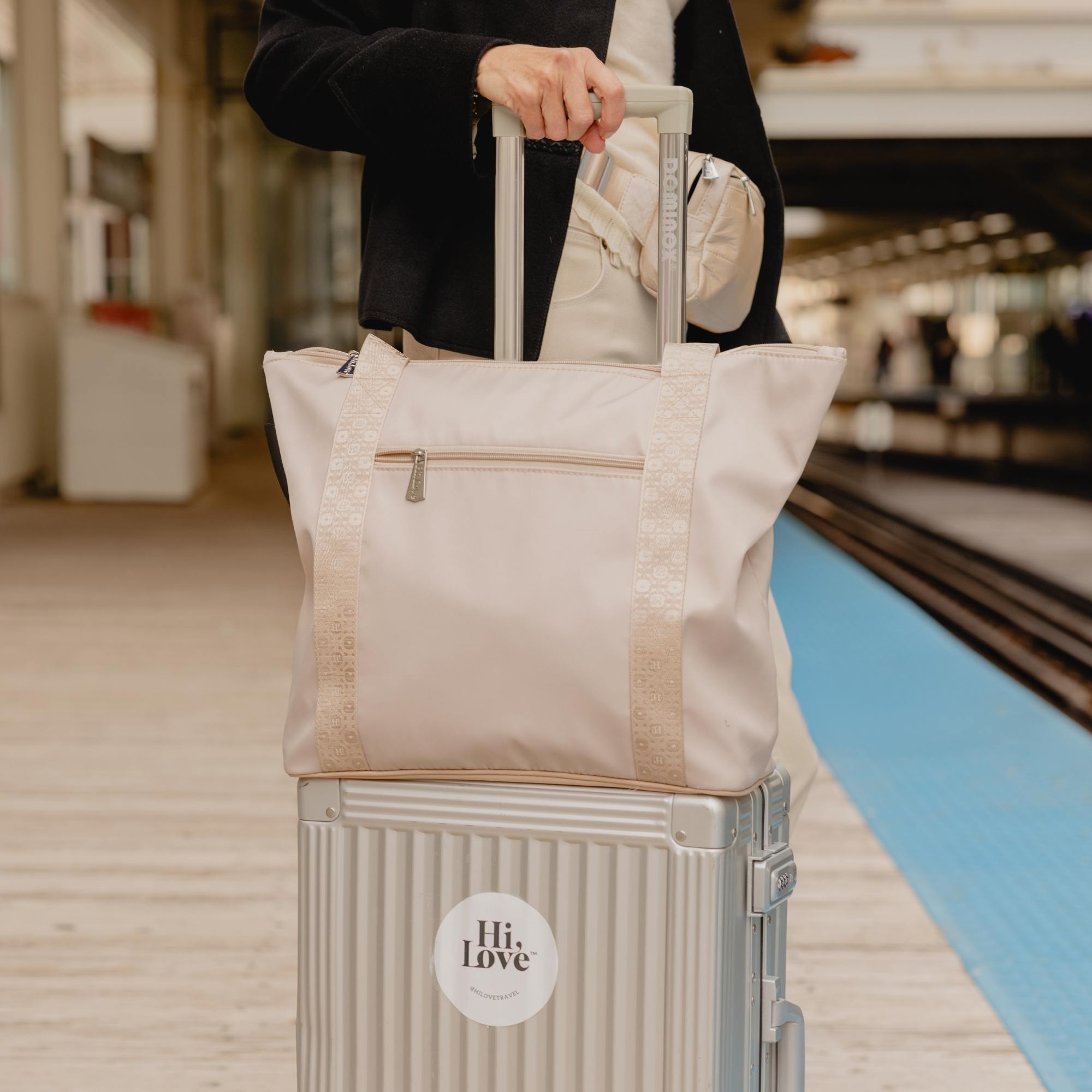 Woman traveling with a beige Signature travel tote on a silver rolling suitcase at a train platform.