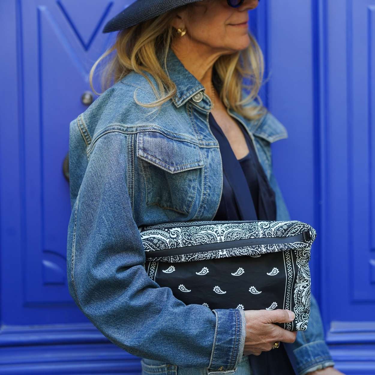 Person wearing a denim jacket holding a black clutch with bandana pattern against a blue background