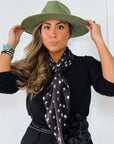 Model wearing the black Signature scarf with white hearts, moons and stars tied at the neck beneath a green hat.
