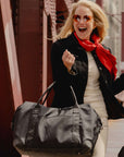 A woman in a black blazer, cream sweater, and bright red scarf carries a black weekender duffel bag while walking outdoors near a bridge structure.
