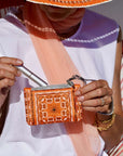 Person holding an orange bandana patterned wallet and a silver lip stick, wearing a white top.