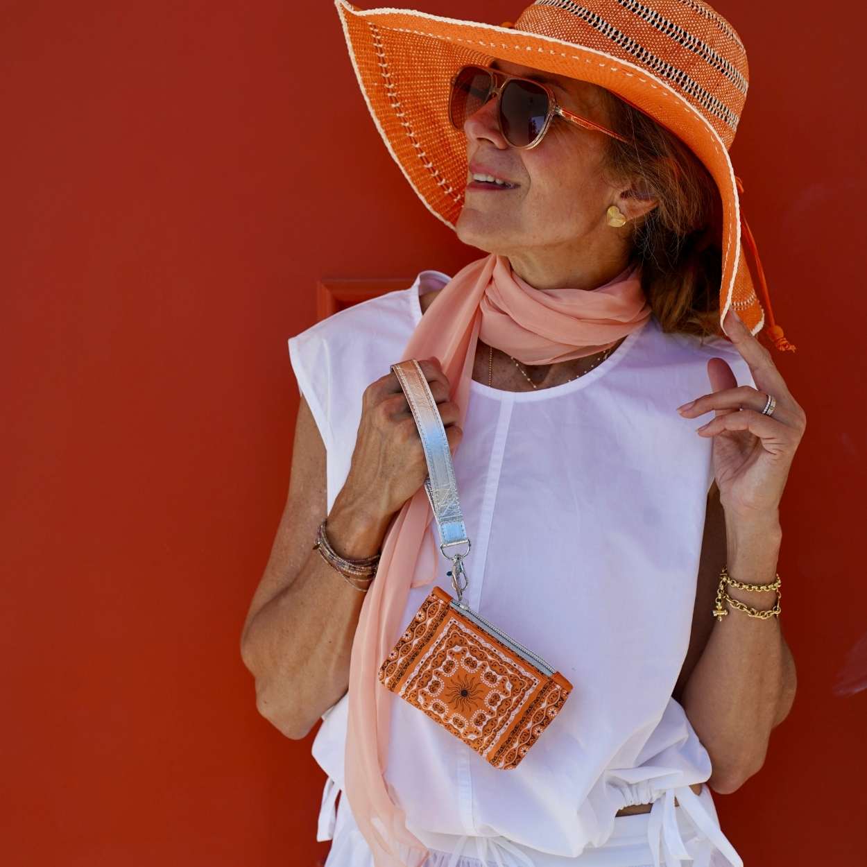 Woman wearing a large orange hat, sunglasses, and holding a orange bandana pouch against a red background