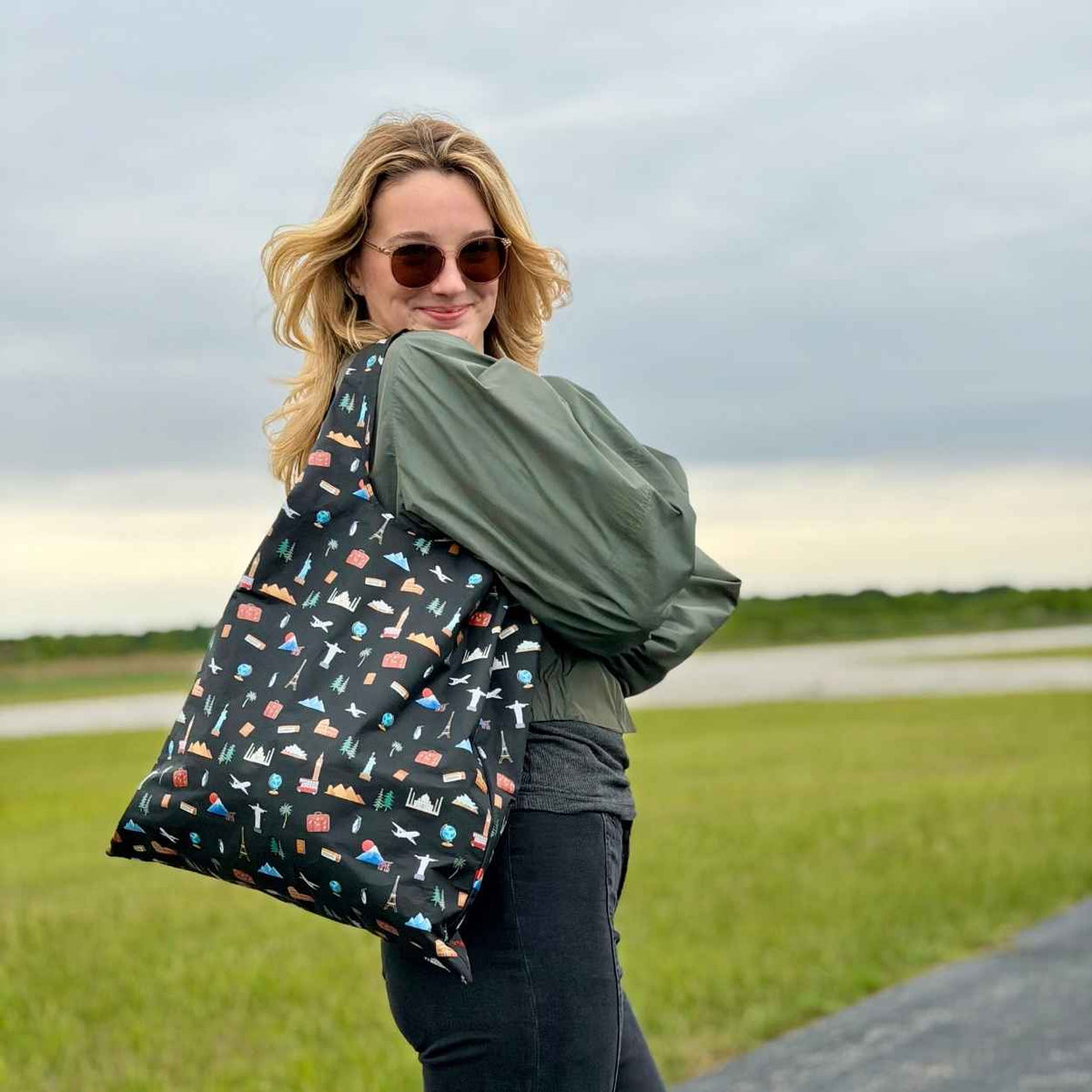 Woman standing in green field wearing army green jacket and black pants, carrying black tote bag with travel inspired print.