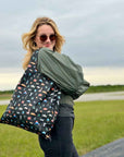 Woman standing in green field wearing army green jacket and black pants, carrying black tote bag with travel inspired print.