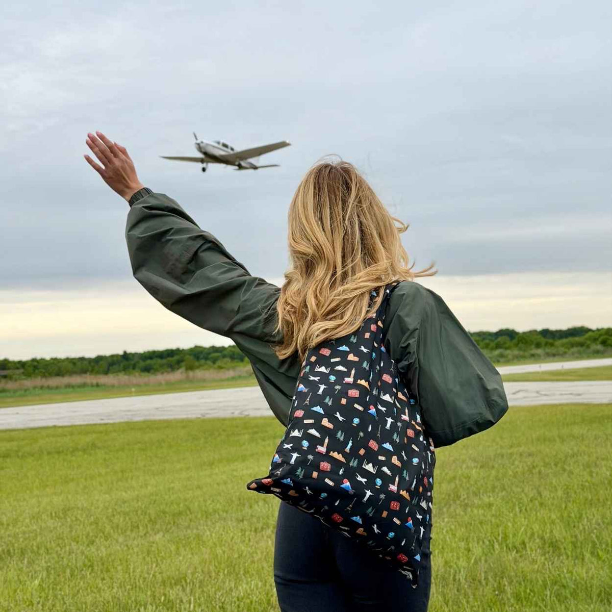 Person standing in field waving at airplane in the sky holding black tote bag with travel inspired pattern of landmarks from the seven continents.