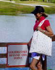 Woman holding white tote bag standing by a sign indicating 'Golfers Only Beyond This Point' near a body of water.