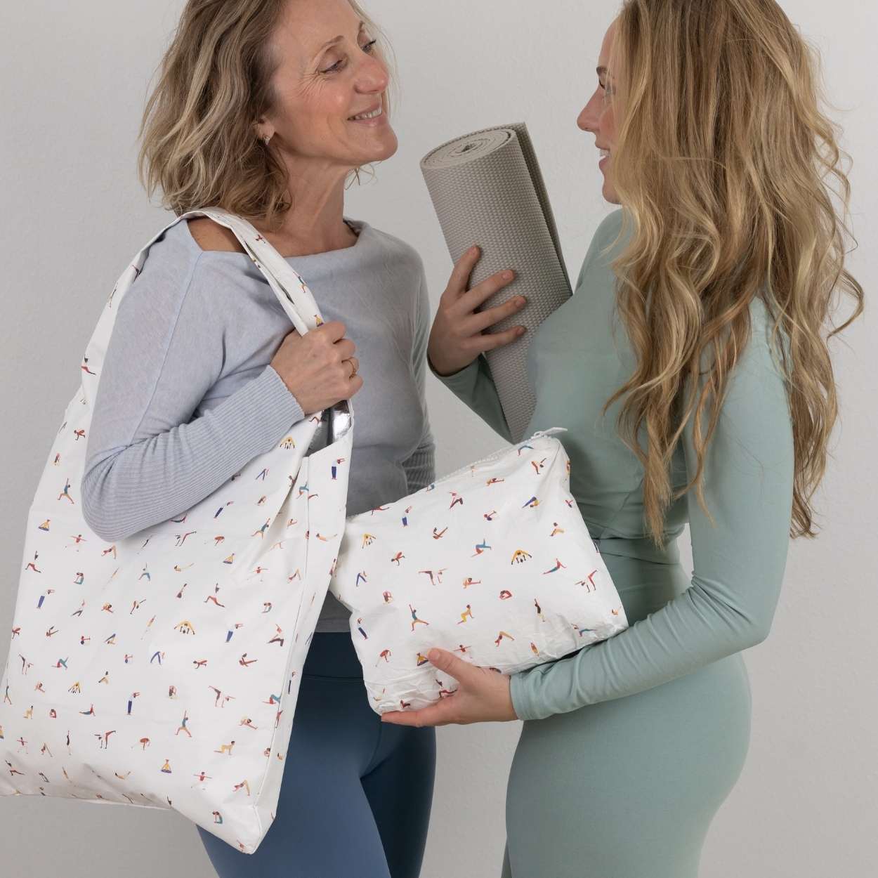 Two women holding yoga patterned bags and a yoga mat against a plain background