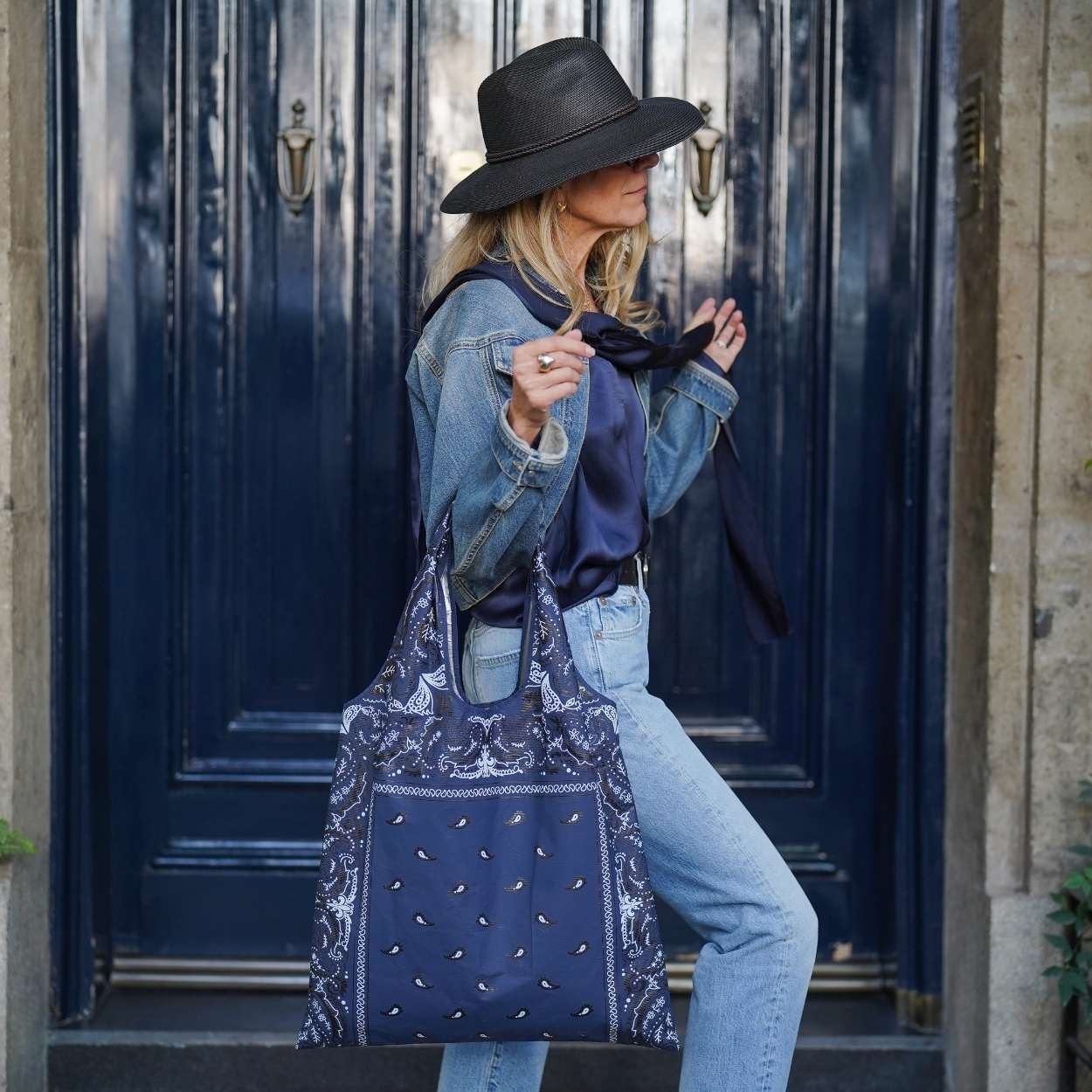 Woman wearing denim outfit and black hat  standing by blue doors holding navy bandana print tote bag 