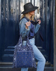 Woman wearing denim outfit and black hat  standing by blue doors holding navy bandana print tote bag 