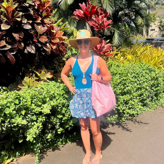 Woman in a blue top and a floral skirt standing in front of colorful plants with a light pink tote bag.