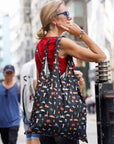 Woman in red top smiling in city setting, carrying black tote bag with a travel landmark inspired pattern.