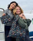 Two women smiling standing on steps of airplane, holding coordinating tote bag and lunchbox with travel landmark inspired print.