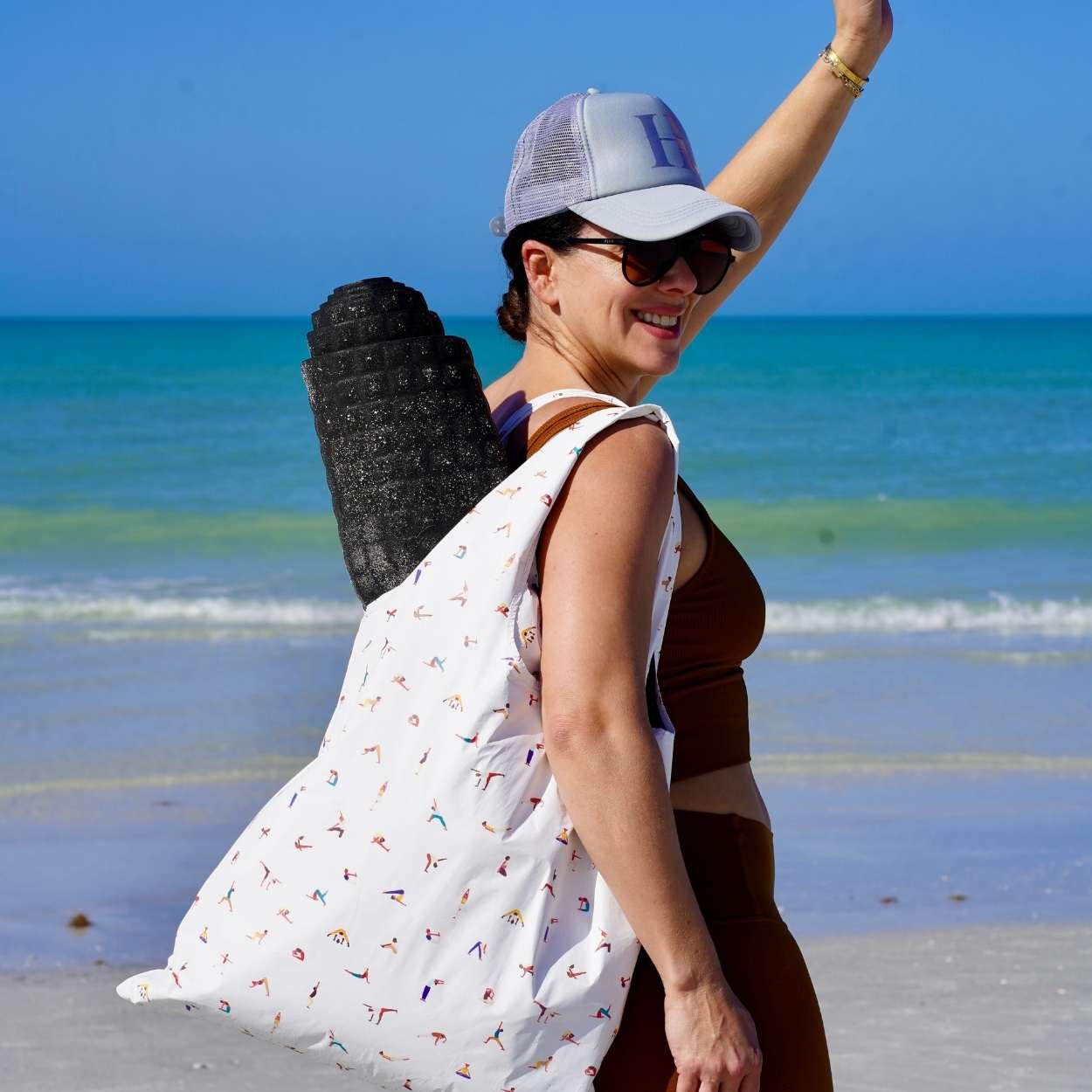 Woman on a beach with a white bag with yoga characters and gray cap, smiling.