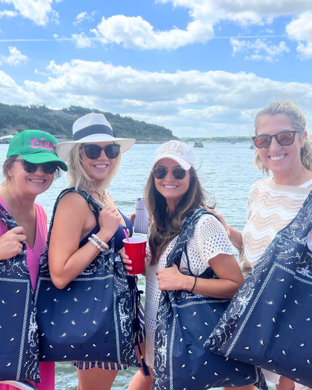 Four women on a boat with a scenic background, wearing navy bandana print carryall totes on their shoulders.