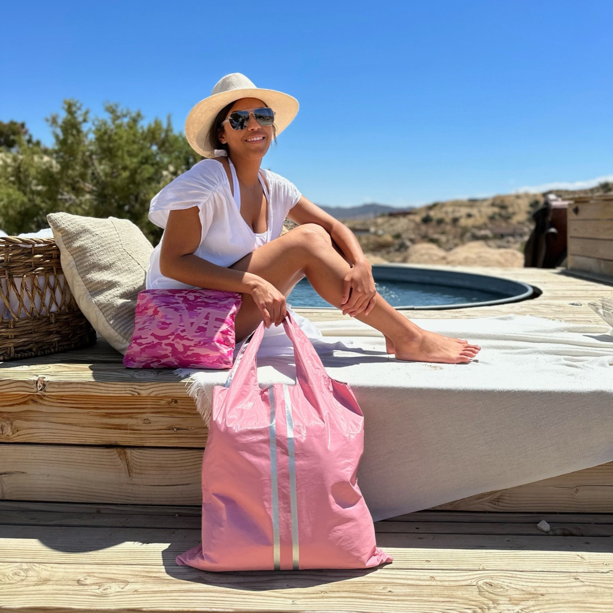 Woman sitting at the edge of the pool with a desert background. She has a pink camo zipper pouch and pink tote bag in front of her. 