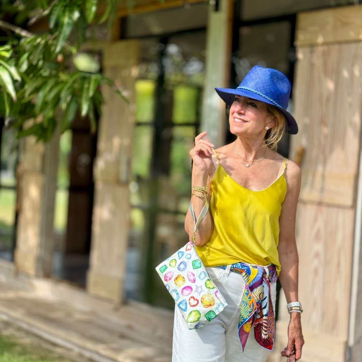 Woman wearing a blue hat and yellow top, holding a colorful bag outdoors.