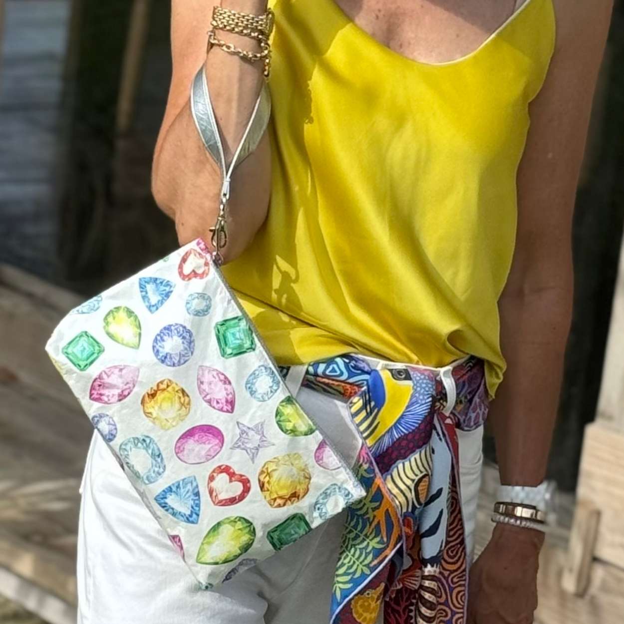 Person holding a colorful clutch with gemstone design, wearing a yellow top and patterned belt.