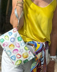 Person holding a colorful clutch with gemstone design, wearing a yellow top and patterned belt.