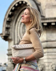 Woman holding a beige clutch bag in an urban setting with architectural elements in the background