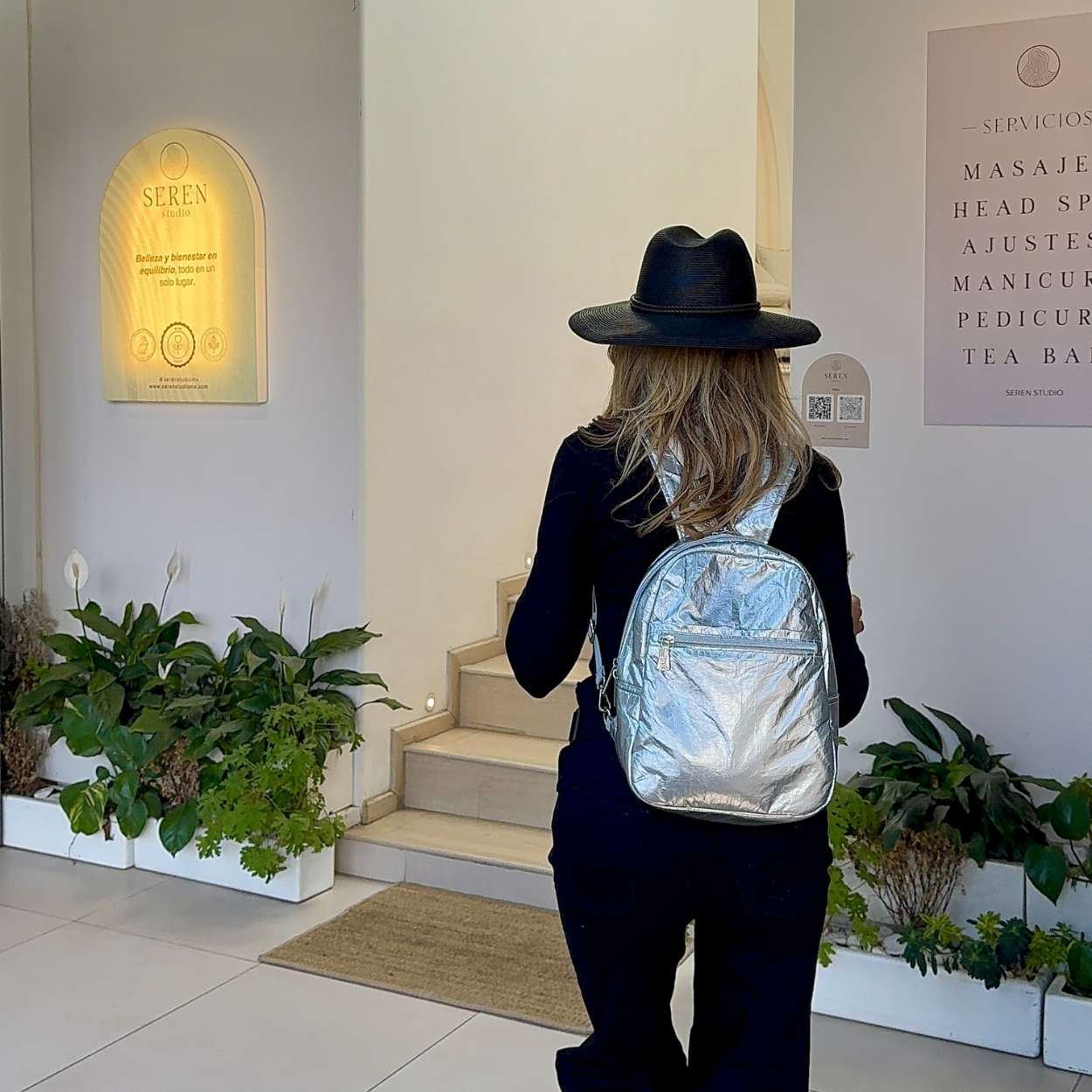 Person wearing a black hat and silver backpack walking into a building with plants and signage.