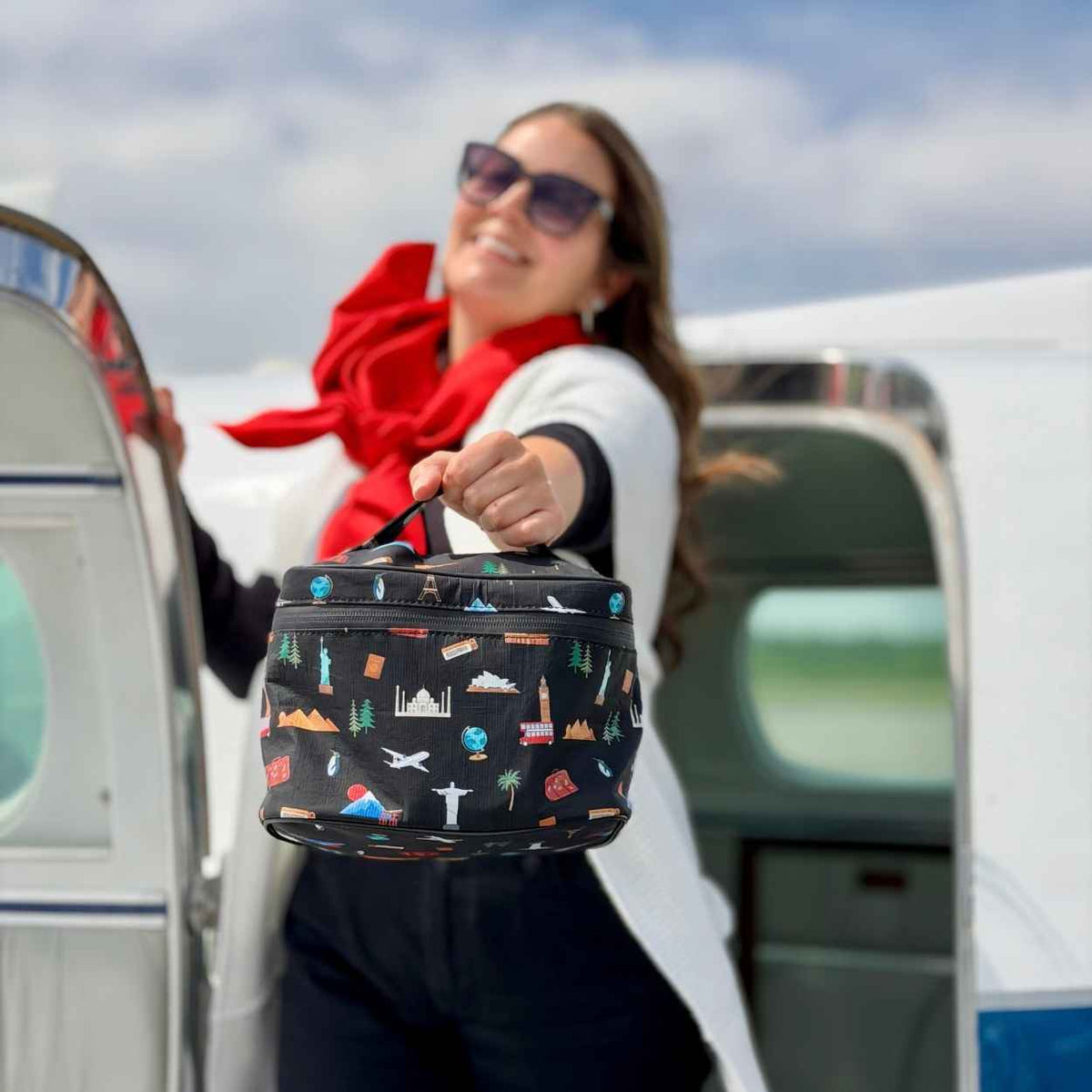 Woman standing on plane in red scarf and sunglasses, smiling holding black travel inspired lunch box.