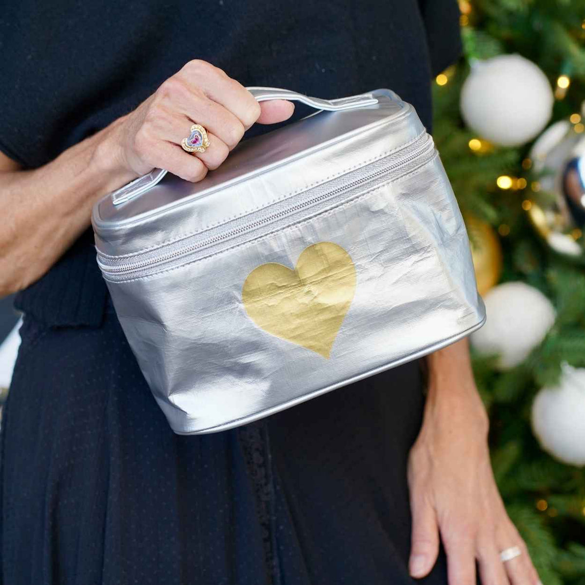 Person holds a metallic silver zippered cosmetic bag with a gold heart on the front. The background shows a blurred Christmas tree with gold ornaments.