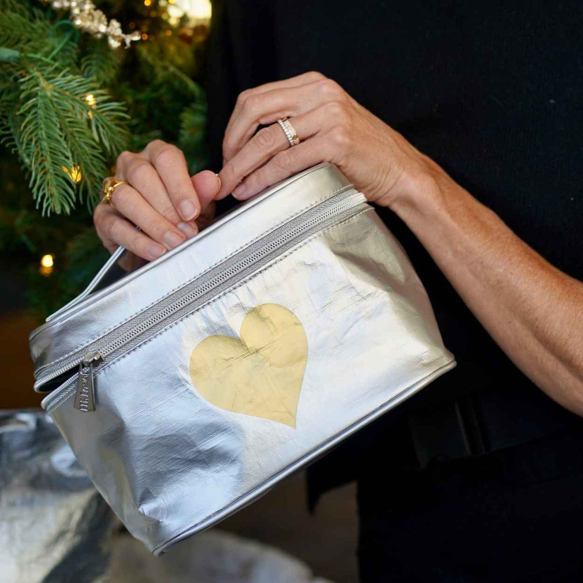 Person holds a metallic silver zippered cosmetic bag with a gold heart on the front. The background shows a blurred Christmas tree with gold ornaments.