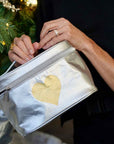 Person holds a metallic silver zippered cosmetic bag with a gold heart on the front. The background shows a blurred Christmas tree with gold ornaments.