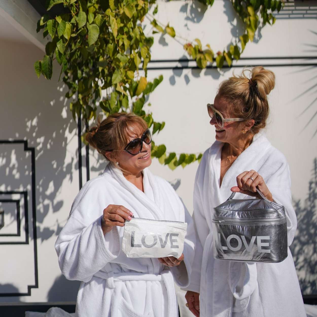 Two women in white robes holding 'LOVE' bags outdoors.