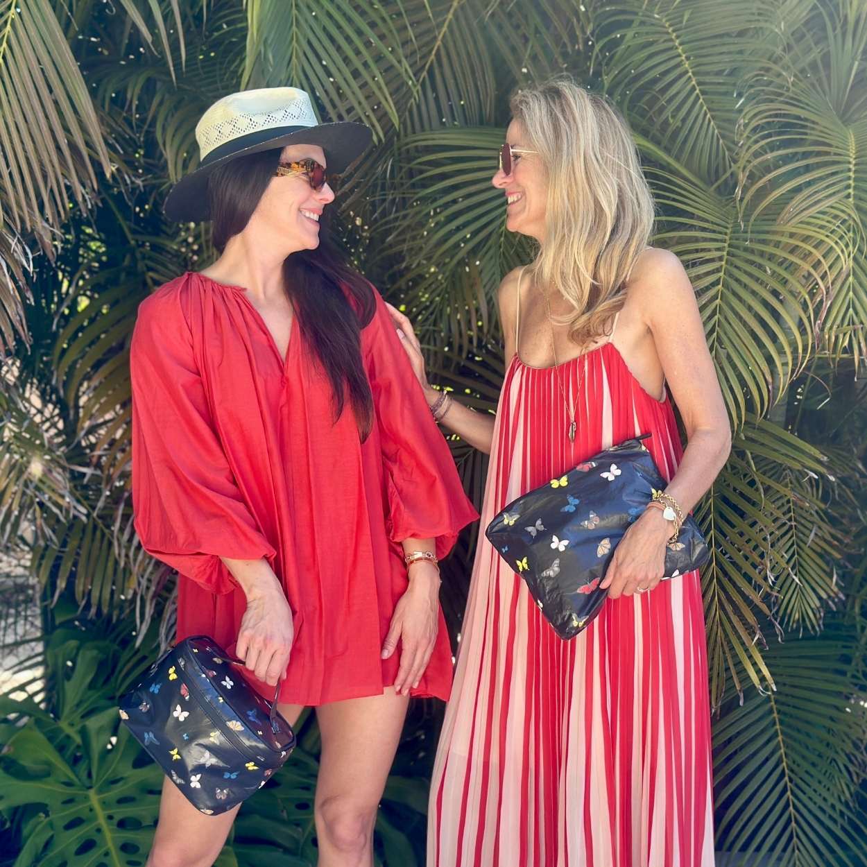 Two women in red outfits standing in front of tropical plants, holding butterfly-patterned bags.
