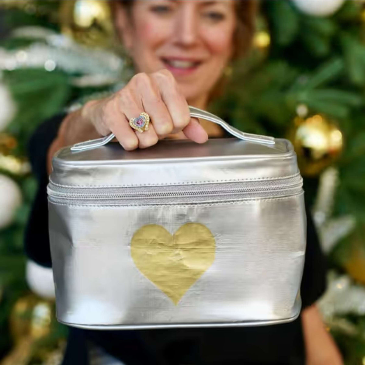 A woman holds a metallic silver zippered cosmetic bag with a gold heart on the front. The background shows a blurred Christmas tree with gold ornaments.