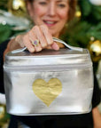 A woman holds a metallic silver zippered cosmetic bag with a gold heart on the front. The background shows a blurred Christmas tree with gold ornaments.