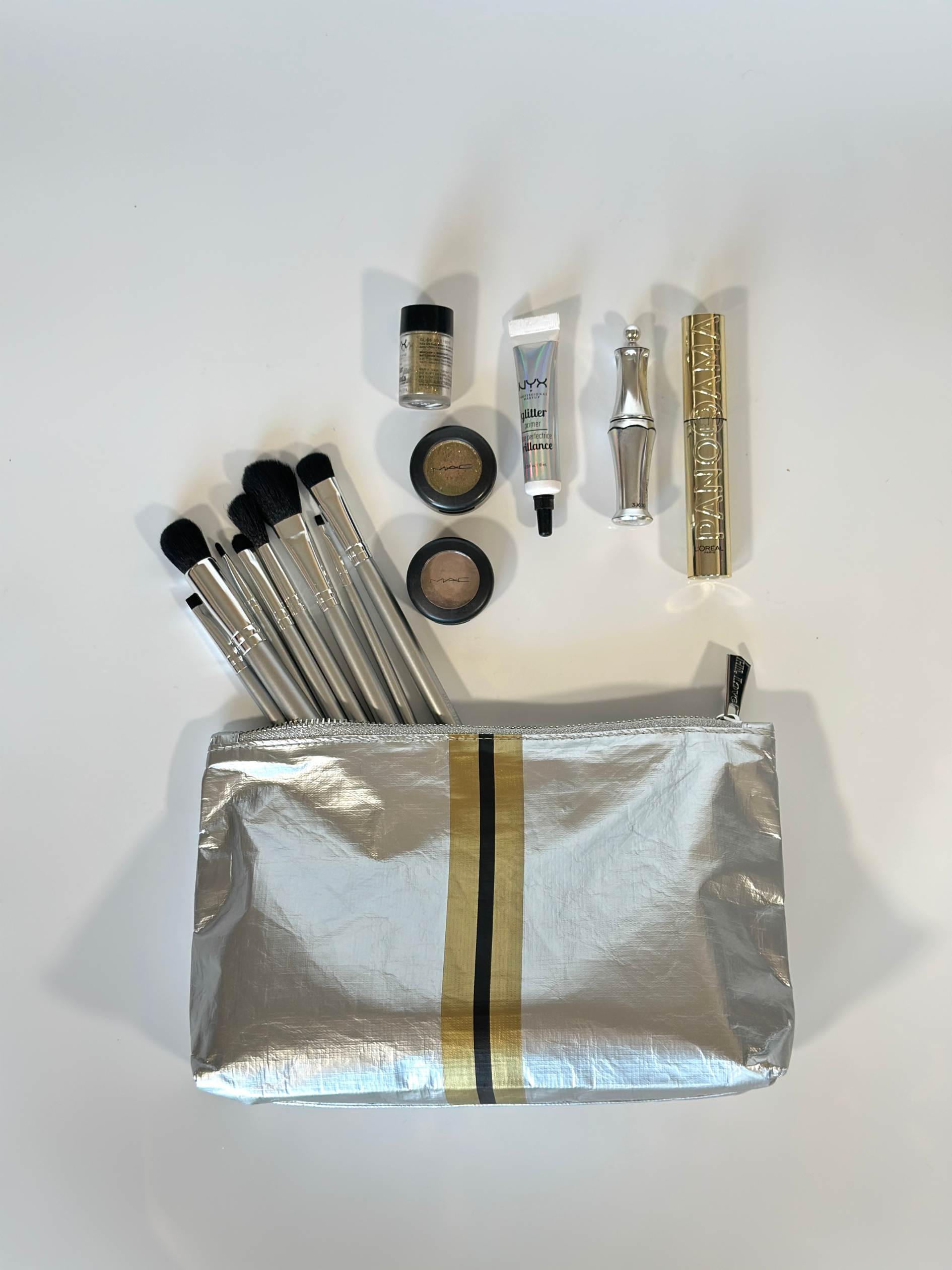 A silver with gold & black stripe cosmetic zipper pouch flat lay image with makeup.