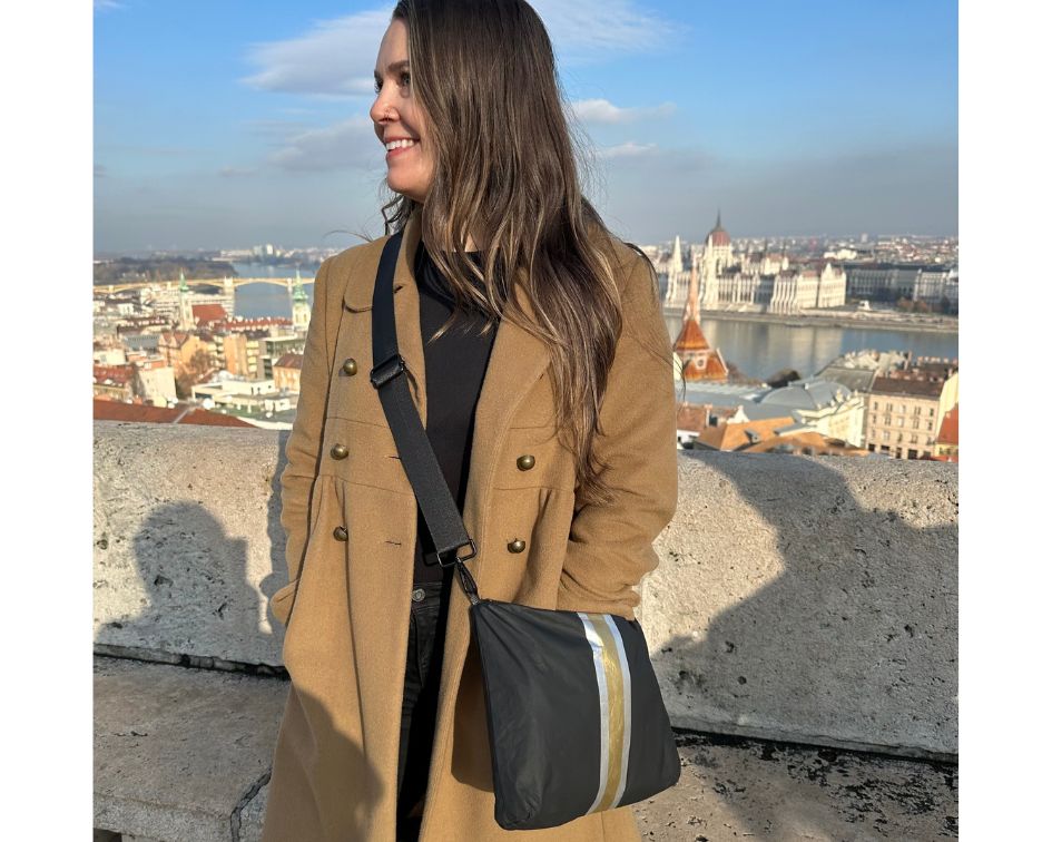 Woman standing overlooking skyline wearing brown peacoat and black crossbody purse with silver and gold stripes