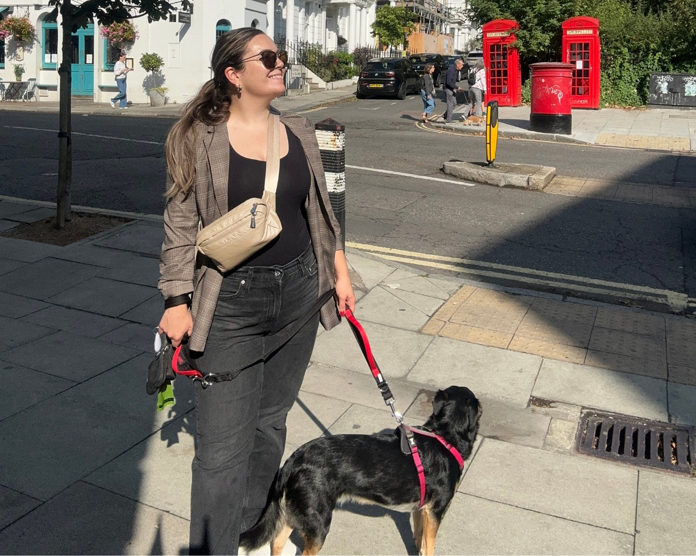 Woman on sidewalk with dog wearing all black and a brown blazer with beige crossbody fanny pack belt bag
