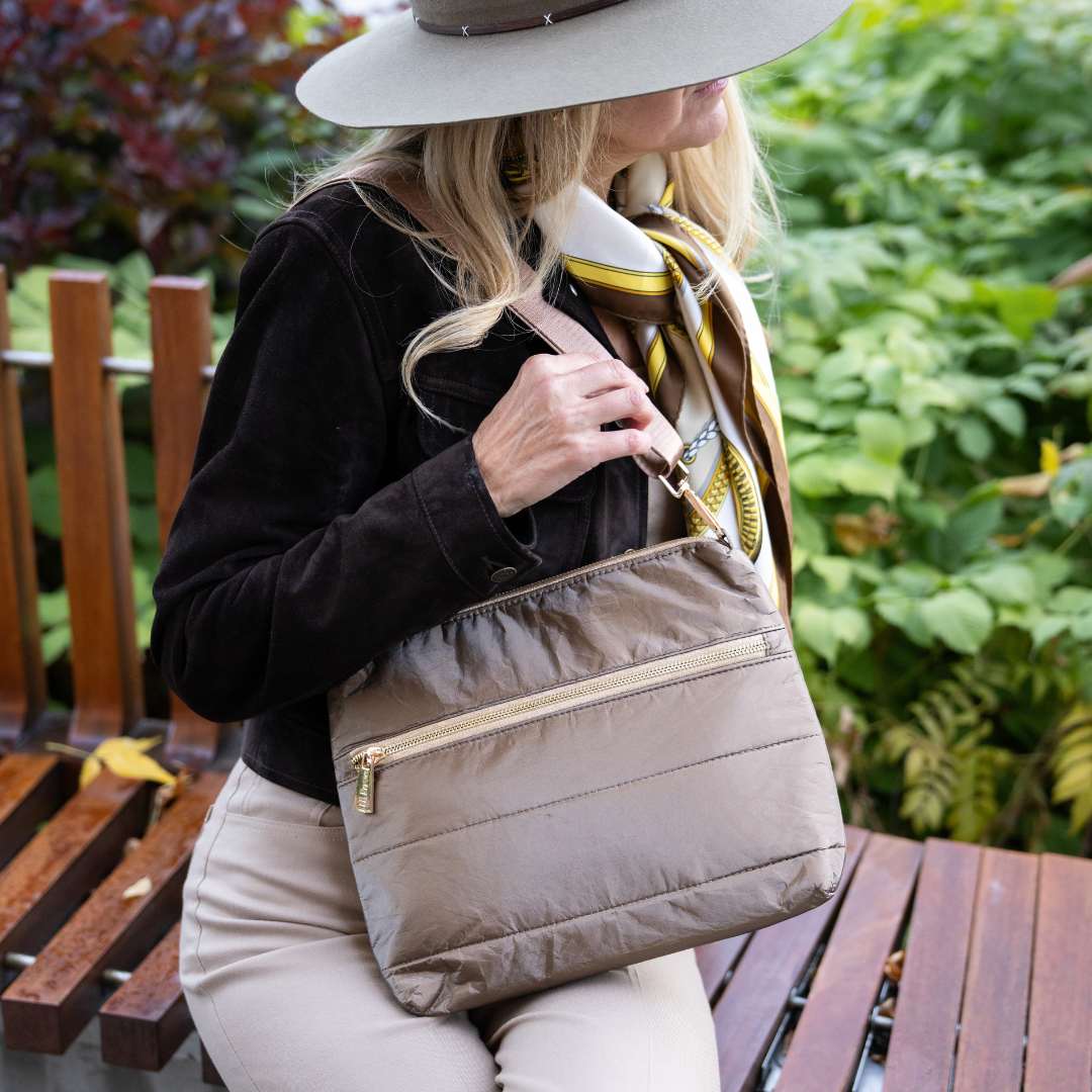 Woman holding a bronze crossody purse outdoors with greenery in the background