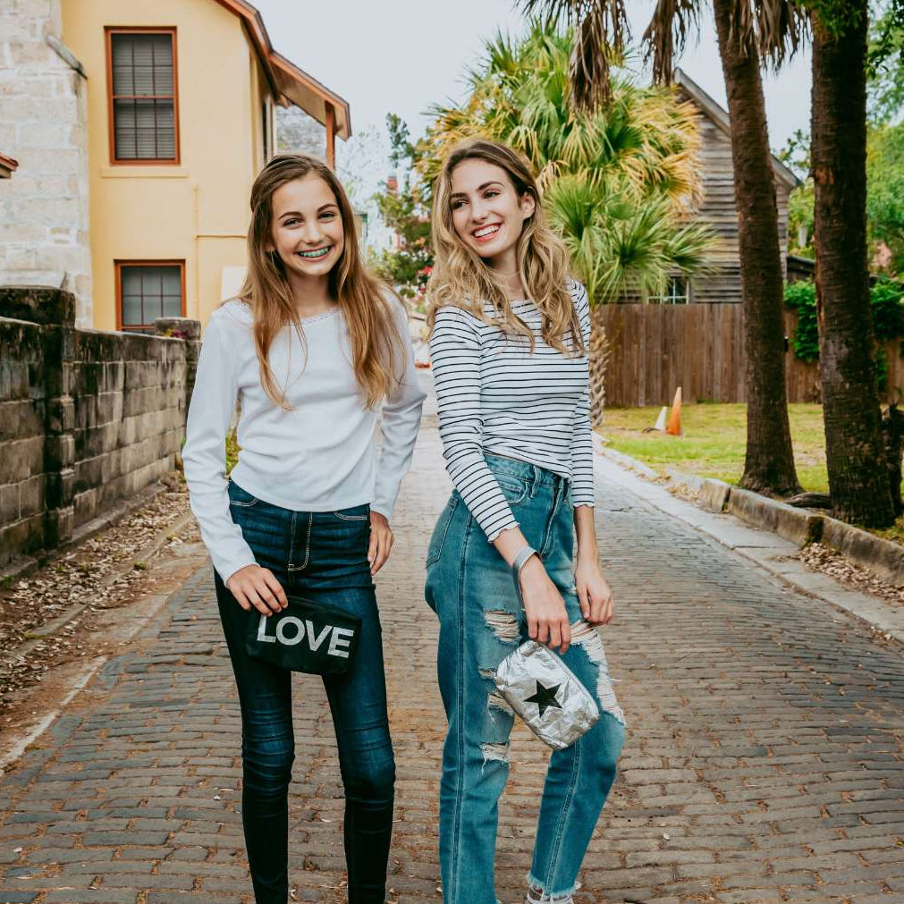 Two women standing on a sidewalk with a house holding black and silver wristlets and palm trees in the background